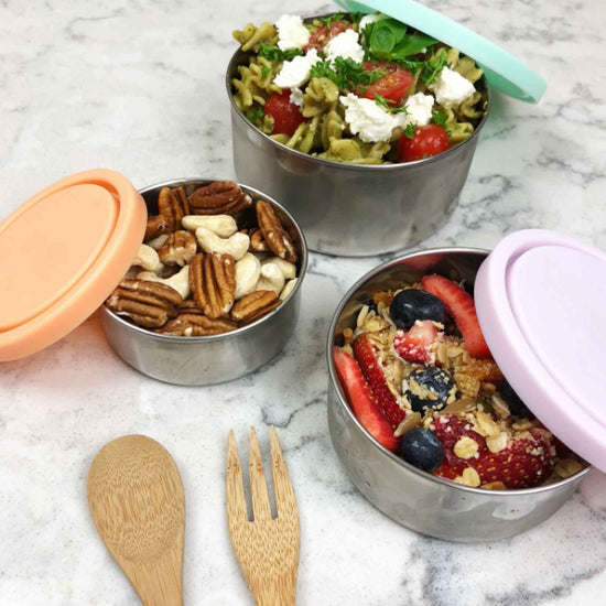 Set of 3 reusable stainless steel food containers. 1 filled with nuts, 1 filled with pasta salad and one with fruit and meusli. Bamboo spoon and fork on the side.