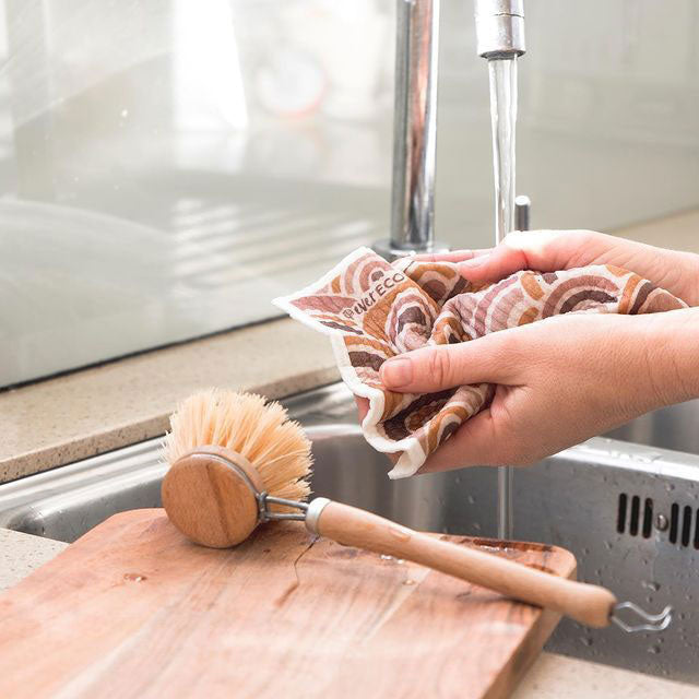 Bamboo handled zero waste dish brush on a wooden chopping board sitting on the sink with a wash cloth in hands. Tap running in the background.