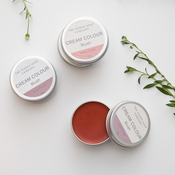 4 Tins of plastic free and toxin free blush. 1 Tin open which is warm red colour. On a white background with some greenery.