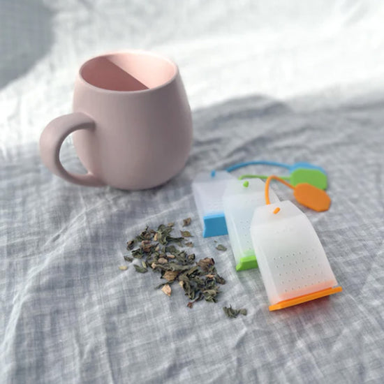 Set of 3 reusable silicone tea bags next to some loose tea leaves and a tea cup. Diminish.